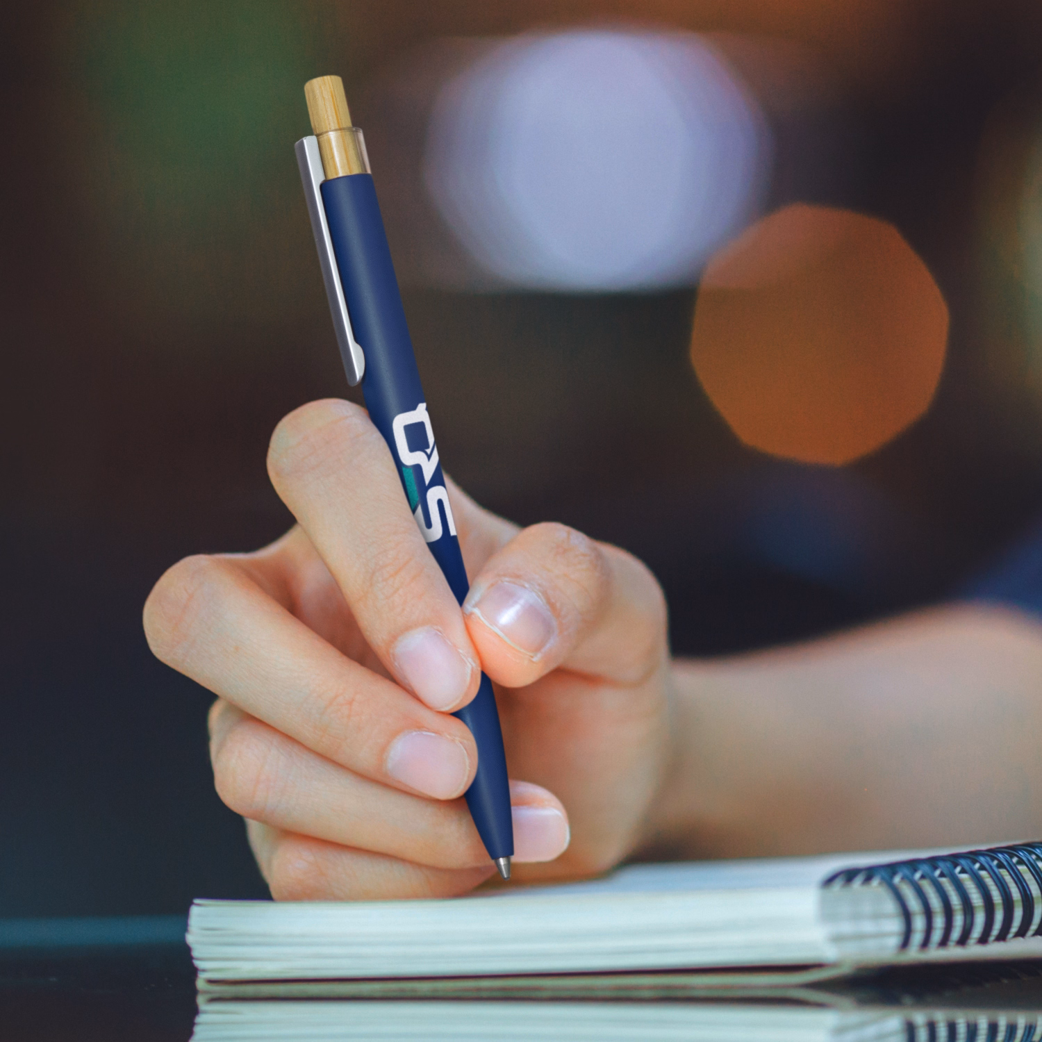 Hand holding branded navy metal pen with bamboo push button, writing in notebook