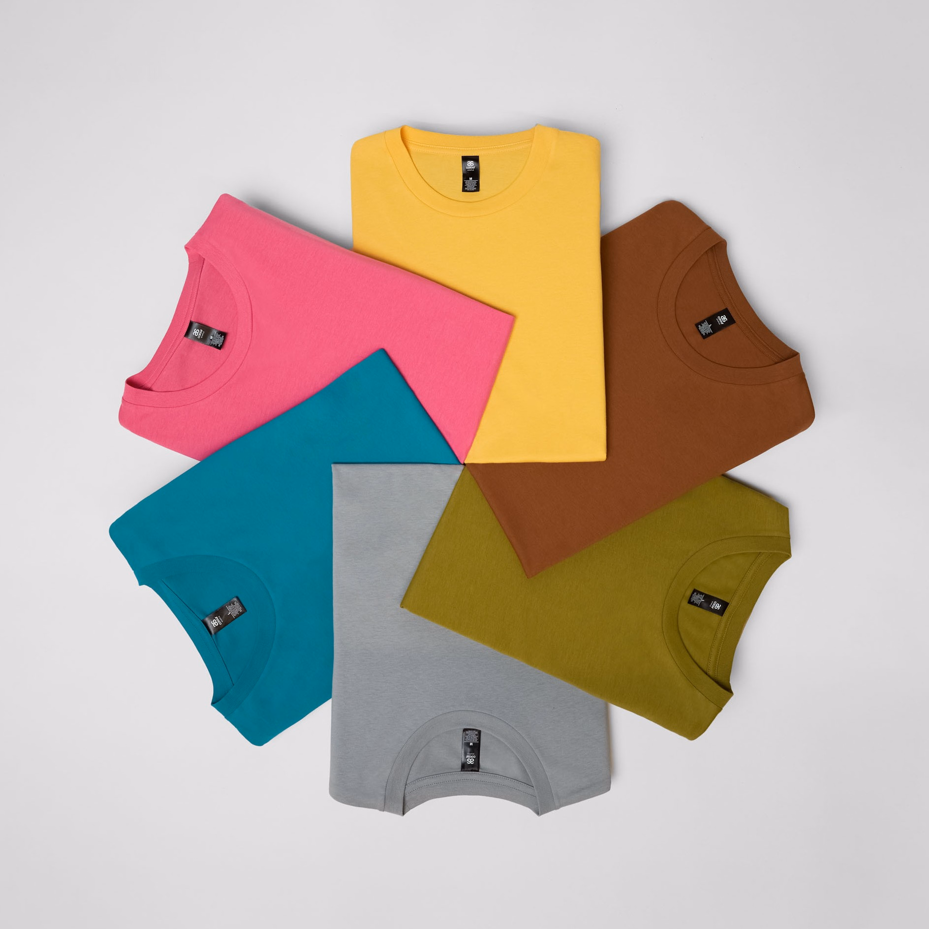 A flat lay of six neatly folded AScolour t-shirts arranged in a circle, showcasing a variety of colours including yellow, pink, teal, grey, brown, and olive green.