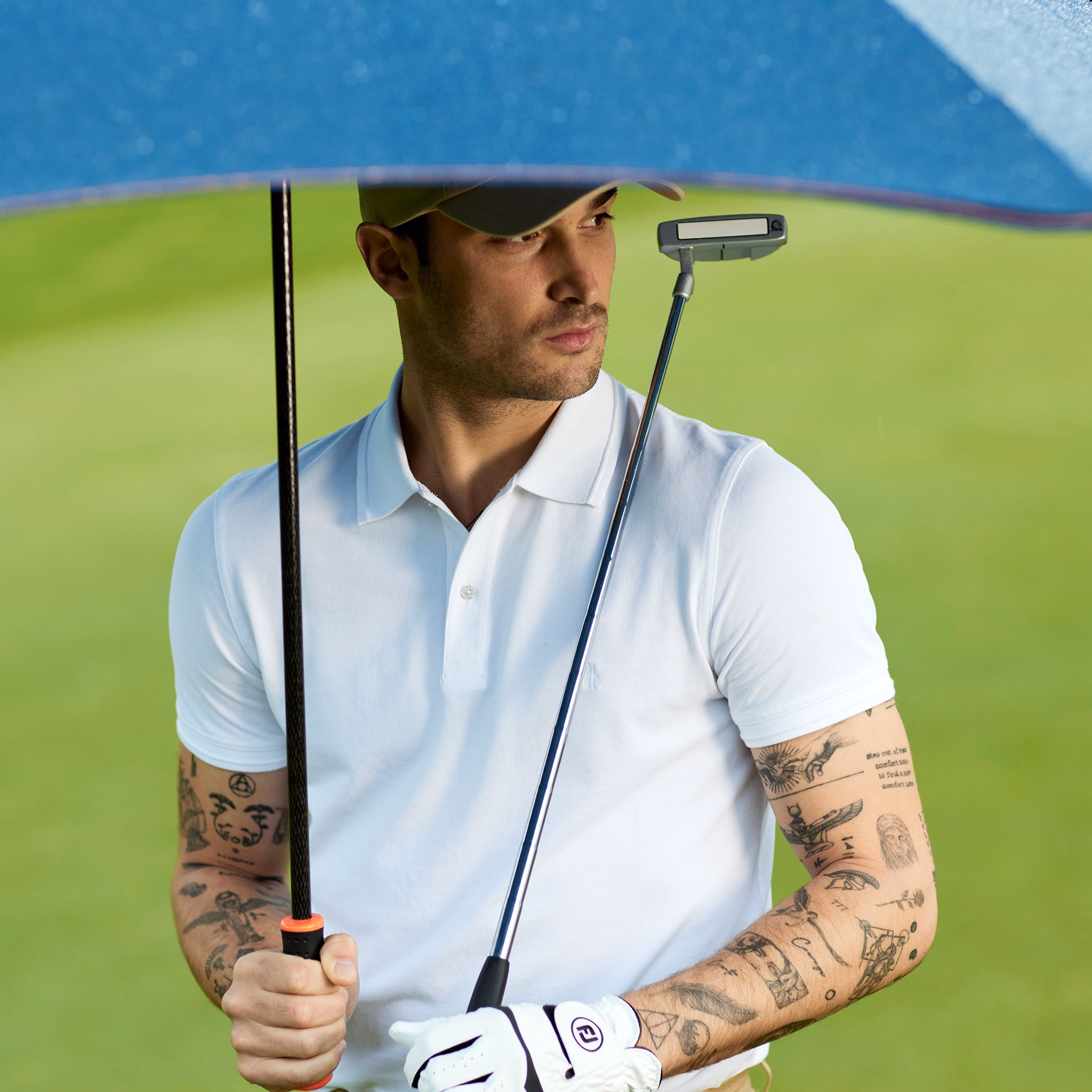 Man standing on golf course holding navy umbrella and golf club while wearing white polo