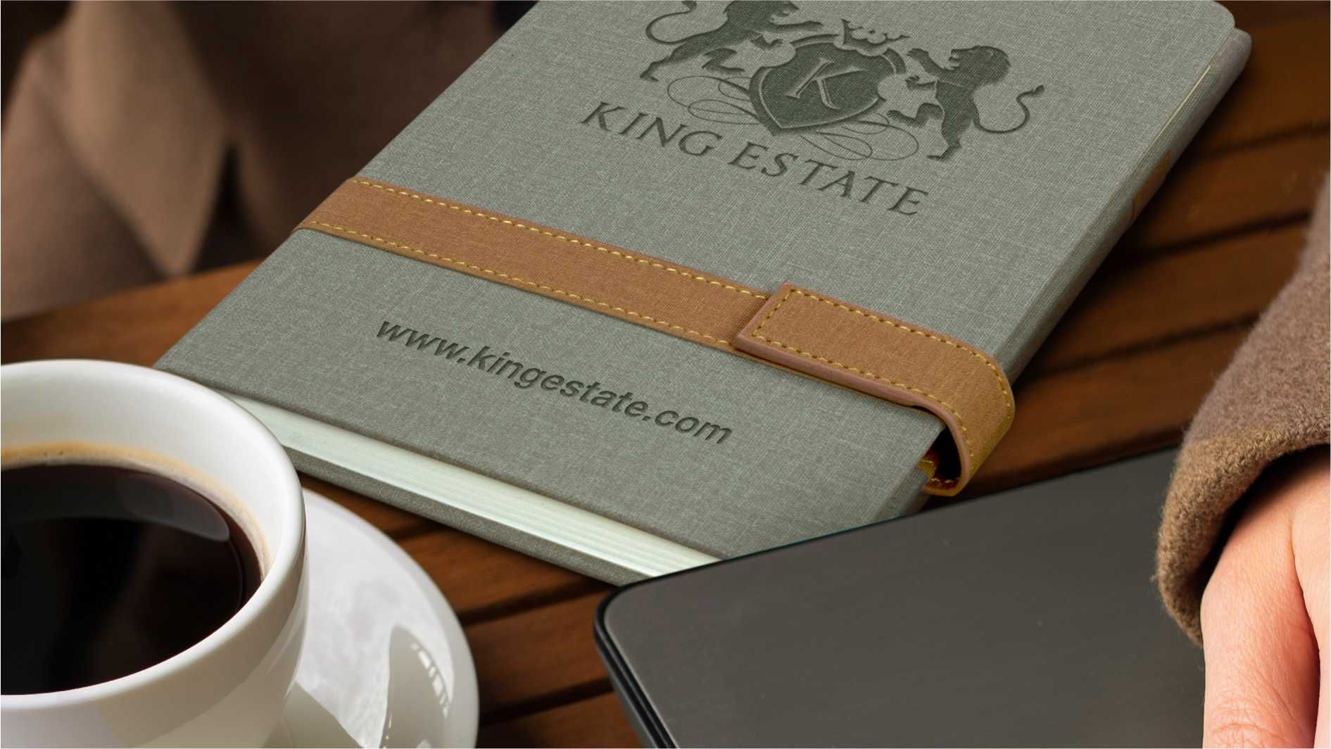 Grey branded notebook with brown strap beside coffee cup and tablet.