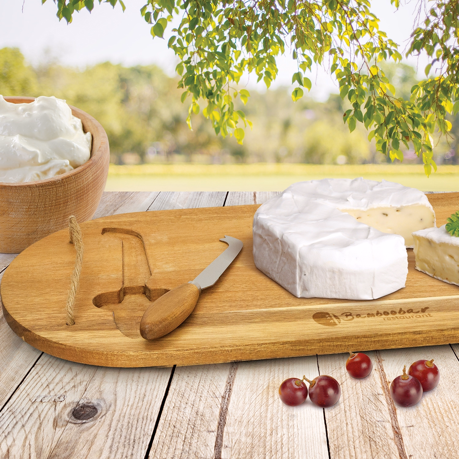 Wooden cheese board with knife, soft cheese, and grapes on outdoor table.