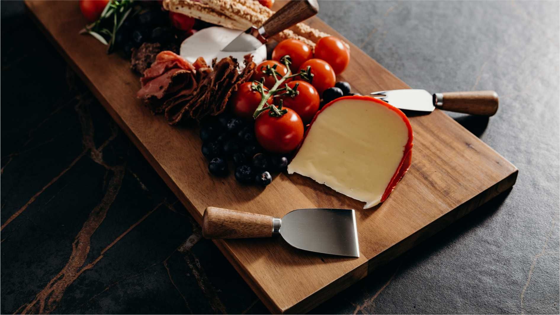 Cheese and antipasto platter on wooden serving board with stainless steel knives.