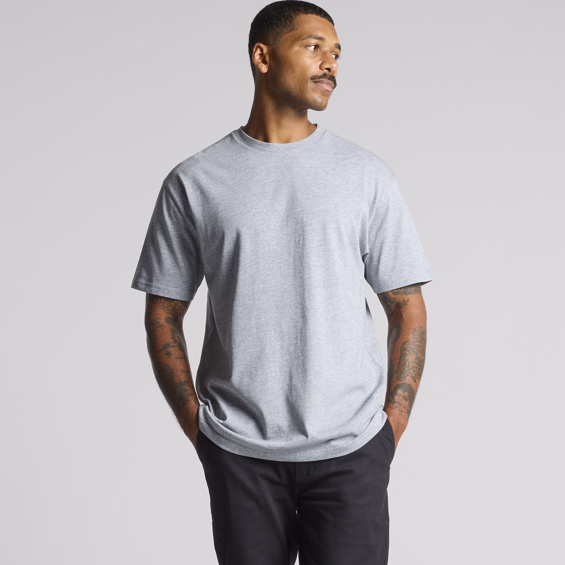 A man wearing a light grey t-shirt and dark trousers, standing with hands in pockets against a plain background, showcasing a relaxed casual fit.