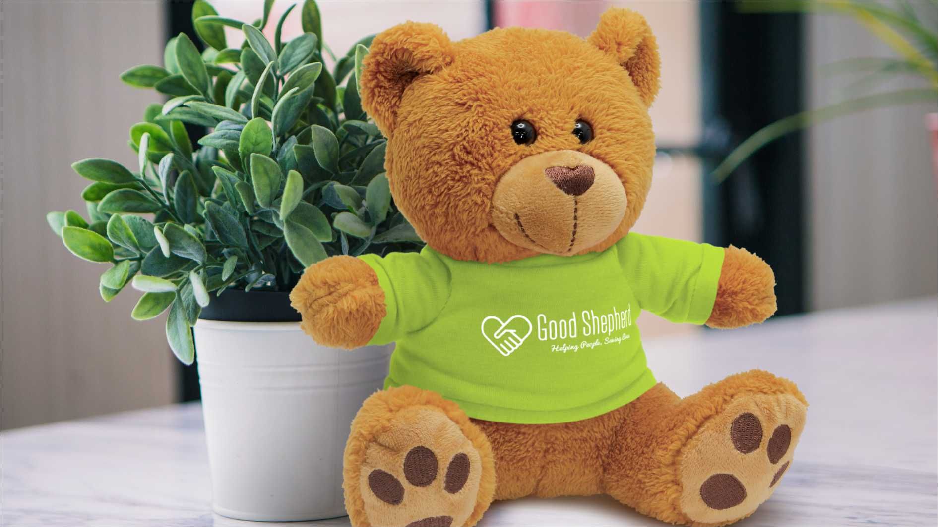 Brown plush teddy bear in green branded T-shirt beside potted plant.