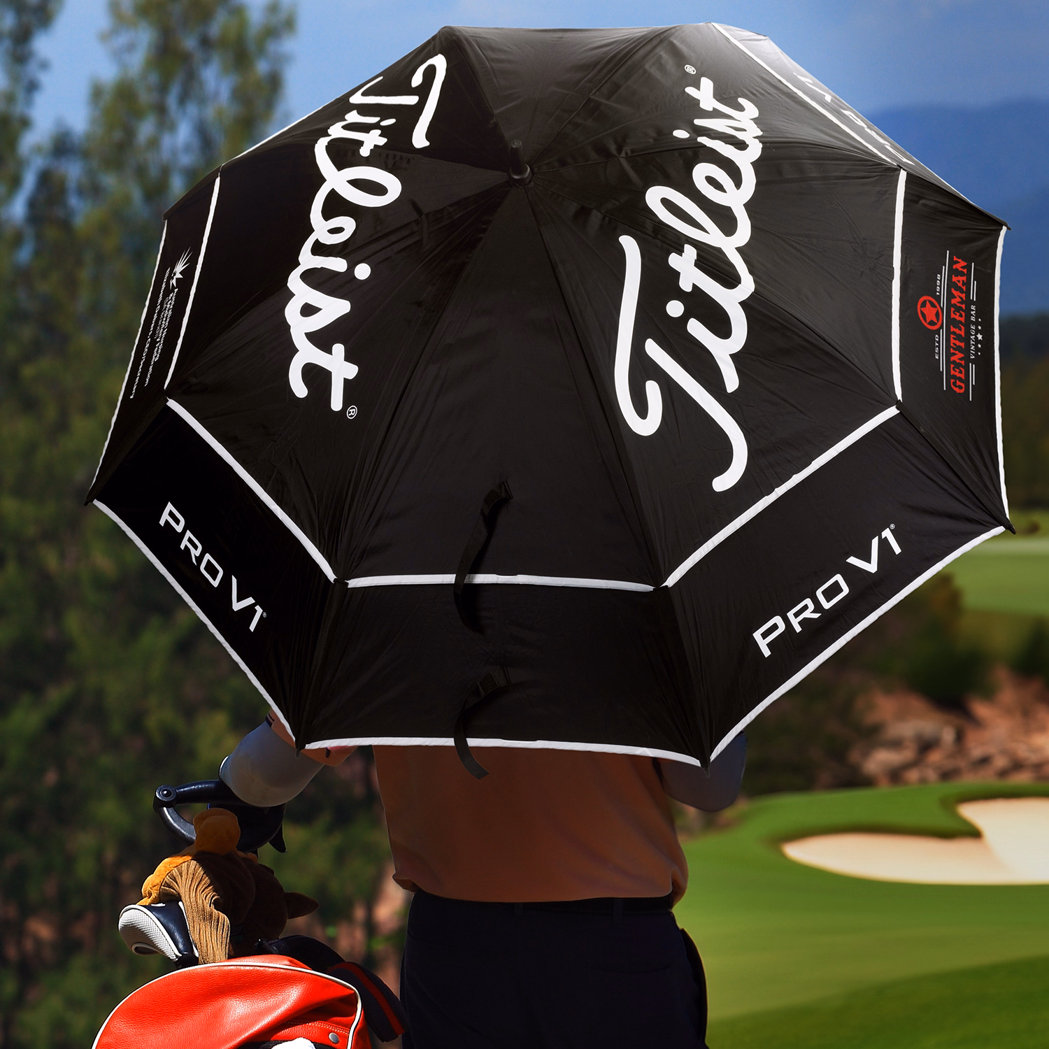 Golfer holding a Titleist Tour Double Canopy Umbrella on a golf course, black canopy with white Titleist and PRO V1 logos