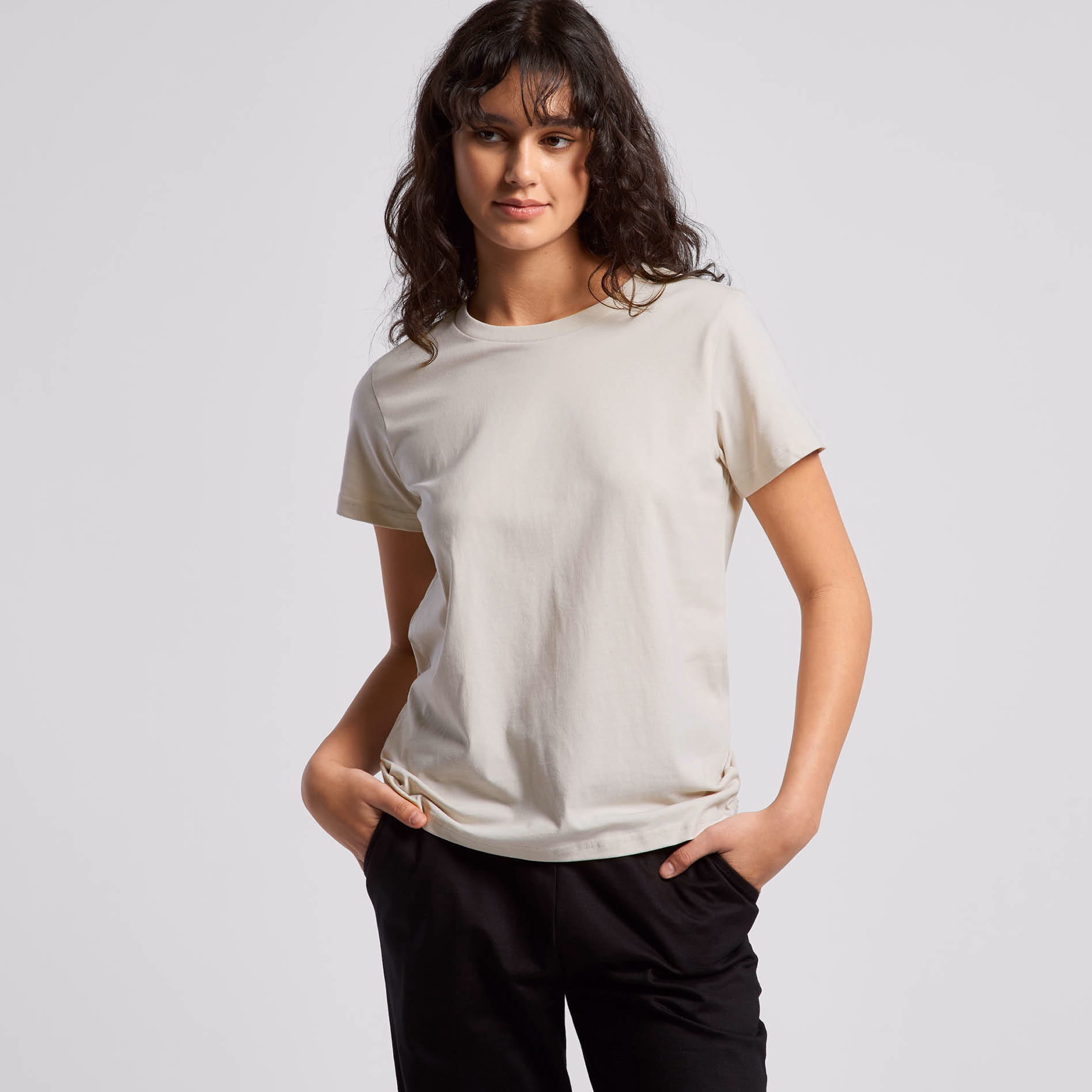 A woman wearing a light beige t-shirt and black pants, standing with hands in pockets against a plain studio background, showcasing a classic women’s casual tee fit.